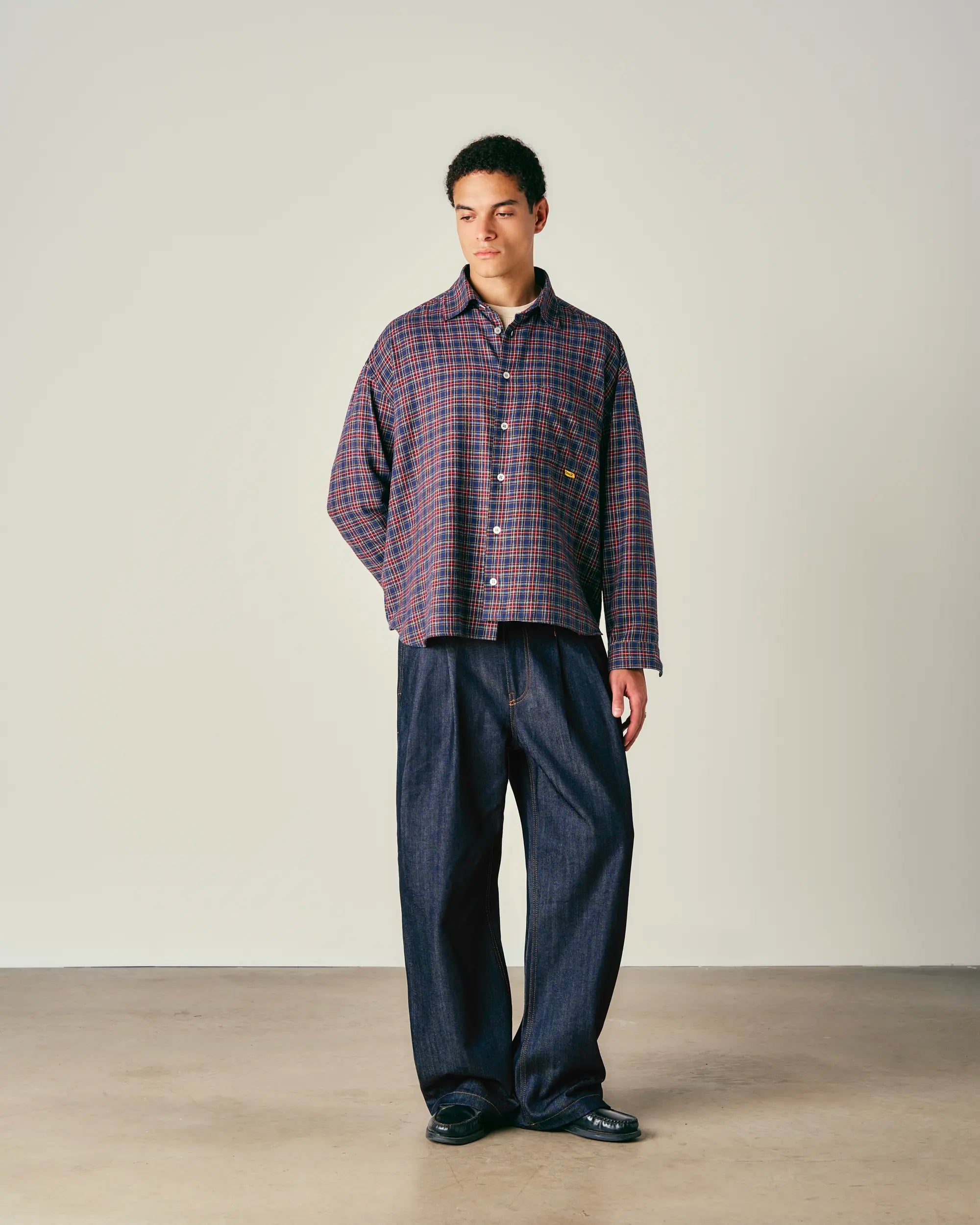 Man wearing a plaid shirt and dark pants against a plain background